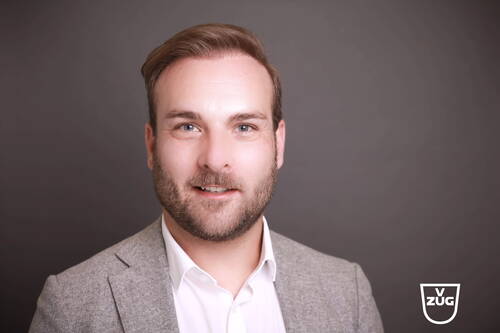 Lächelnder Mann im grauen Blazer und weißem Hemd vor einfarbig dunkelgrauem Hintergrund mit V-ZUG-Logo unten rechts
