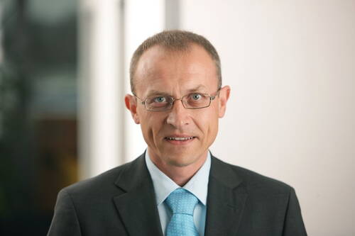 Geschäftsmann mittleren Alters mit Brille und blauem Anzug, der in heller Büro-Umgebung direkt in die Kamera lächelt