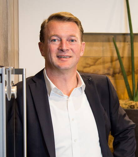 Mittelalterlicher Mann im Anzug lehnt lächelnd an einer Glastür in einem modernen Büro mit Holzwand und hohen Grünpflanzen im Hintergrund