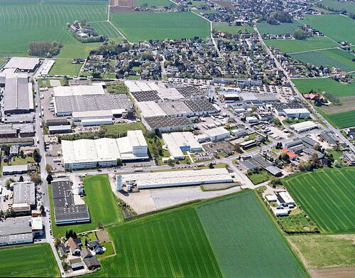 Luftaufnahme eines weitläufigen Industriegebiets mit großen Hallen, umgeben von grünen Feldern und angrenzender Wohnsiedlung im Hintergrund