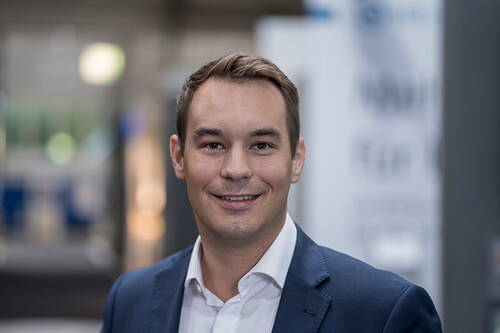 Junger Mann im Business-Anzug lächelt direkt in die Kamera in einer modernen Büroumgebung mit unscharfem Hintergrund