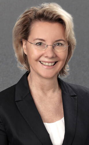 Lächelnde Frau mittleren Alters mit kurzem blondem Haar, Brille und dunklem Blazer vor neutral grauem Hintergrund