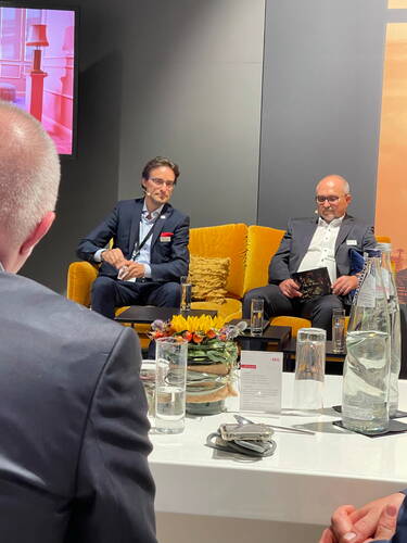 Zwei Geschäftsmänner im Anzug diskutieren mit Headset-Mikrofonen auf einem gelben Sofa, während vor ihnen auf einem Konferenztisch Wasserflaschen, Gläser und ein Blumenarrangement stehen.