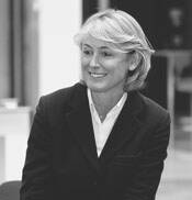 Lächelnde Frau mit kurzem blondem Haar im dunklen Blazer sitzt in einem Innenraum und blickt seitlich aus dem Bild.
