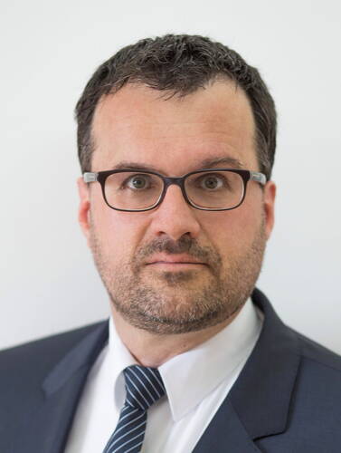 Mittelalterlicher Mann mit kurzer dunkler Frisur, Brille und Dreitagebart im Anzug und gestreifter Krawatte blickt ernst in die Kamera vor neutralem Hintergrund
