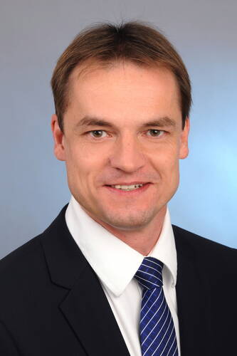 Lächelnder Mann im Anzug mit weißem Hemd und blau gestreifter Krawatte vor neutral blau-grauem Hintergrund