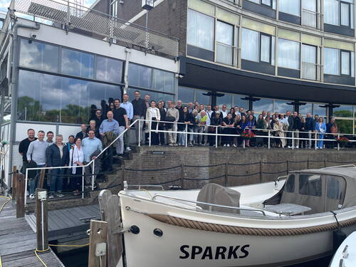 Große Geschäftsgruppe posiert lächelnd auf einer Terrasse am Wasser vor einem modernen Gebäude, während im Vordergrund mehrere festgemachte Boote liegen.
