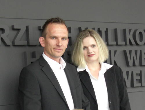 Zwei Geschäftsleute in schwarzen Anzügen stehen nebeneinander vor einer grauen Wand mit teils lesbarem Schriftzug.