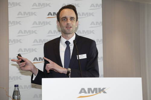 Geschäftsmann im Anzug hält eine Präsentation am Rednerpult und gestikuliert mit einem Presenter vor einer AMK-Hintergrundwand