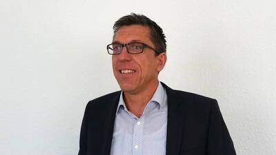 Mittelalterlicher Mann mit Brille lächelt im Anzug vor einer weißen Wand und blickt leicht nach links.