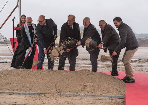 Mehrere festlich gekleidete Männer heben beim offiziellen Spatenstich einer Baustelle gleichzeitig Sand mit Schaufeln aus einem Erdhaufen, während eine junge Frau mit Mikrofon im Hintergrund zusieht.