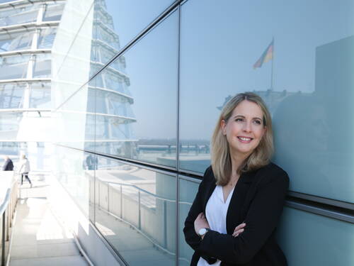 Lächelnde Businessfrau lehnt mit verschränkten Armen an einer Glasfassade auf der Dachterrasse des Reichstagsgebäudes, im Hintergrund spiegelt sich die deutsche Flagge im Glas.