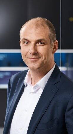 Mittelalterlicher Mann im Businessanzug lächelt freundlich in die Kamera und steht vor einem modernen, verschwommenen Bürohintergrund