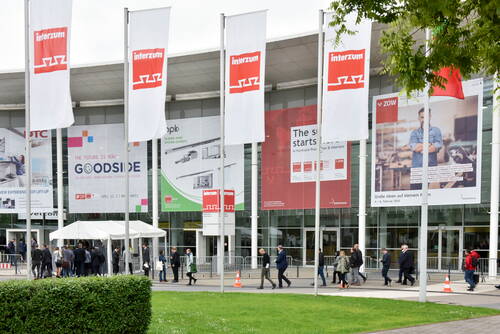 Besucher strömen durch den Eingangsbereich der Messe interzum, gesäumt von weißen Fahnen und großen Werbebannern an der Glasfassade.