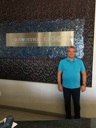 Mann in türkisfarbenem Polohemd steht lächelnd vor einer Wandinstallation mit dem Metalllogo von Sub-Zero und Wolf in einem modernen Empfangsbereich