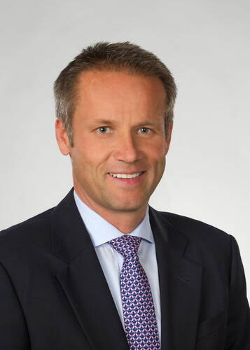 Mittelalterlicher Mann im dunklen Geschäftsanzug und gemusterter Krawatte lächelt freundlich in einem professionellen Porträt vor neutral grauem Hintergrund.
