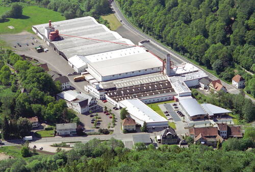 Luftaufnahme eines großflächigen Industrie- und Lagerkomplexes mit hellen Hallendächern, umgeben von Straßen, Wohnhäusern und dicht bewaldeten Grünflächen.
