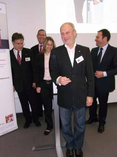 Älterer Mann im gestreiften Sakko spricht lächelnd am Rednerpult, während vier Geschäftsleute im Hintergrund in Anzügen bei einer Firmenveranstaltung stehen und lachen.
