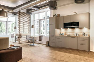 Helle, moderne Büroküche mit beiger Einbauzeile, großen Fenstern, sichtbaren Lüftungsrohren und angrenzendem Sitzbereich mit kleinen Tischen und Stühlen