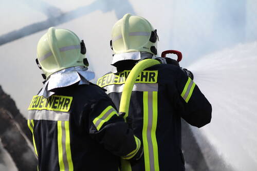 Zwei Feuerwehrleute in Schutzkleidung richten einen kräftigen Wasserstrahl aus einem Schlauch auf einen Brandherd.