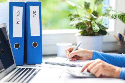 Person berechnet am Schreibtisch Strom- und Gaskosten mit Taschenrechner, Notizblock und blauen Ordnern für Energieverträge neben dem Laptop.