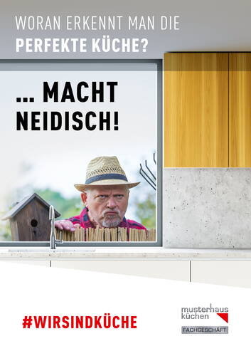 Älterer Mann mit Strohhut blickt missgelaunt über den Gartenzaun in eine moderne Küche mit großem Fenster und Holzfronten