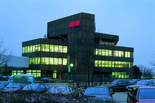 Mehrstöckiges Bürogebäude mit leuchtend grünen Innenräumen und rotem ALNO-Schriftzug in der Dämmerung, umgeben von voll besetztem Parkplatz.