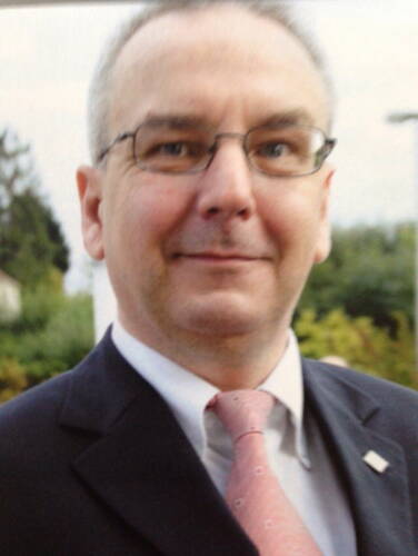 Älterer Mann mit Brille im Anzug und rosafarbener Krawatte lächelt leicht in die Kamera vor einem unscharfen Hintergrund im Freien.