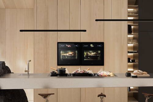 Modern gestaltete Küche mit heller Holzverkleidung, zwei eingebauten Hightech-Backöfen und einer Inselarbeitsplatte, auf der Töpfe, Gemüse und frisches Brot für die Zubereitung bereitstehen.