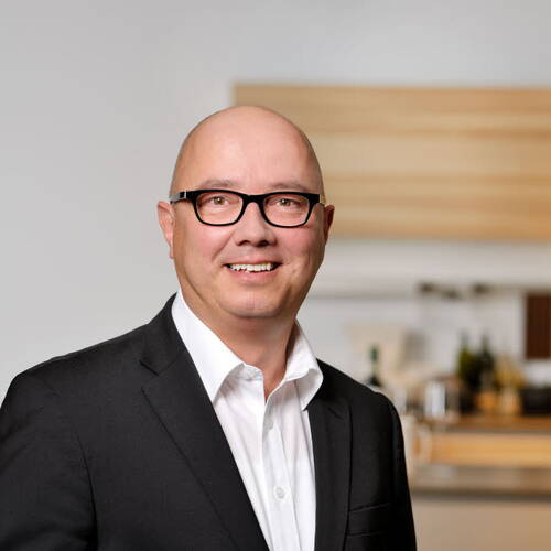 Lächelnder, glatzköpfiger Mann mit Brille in schwarzem Anzug und weißem Hemd steht in einer modern eingerichteten Küche.