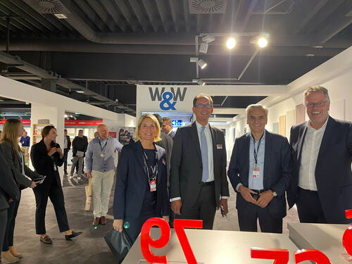 Gruppe von Geschäftsleuten in Anzügen lächelt in einer modernen Messehalle unter dem Schriftzug „W&W“ in die Kamera