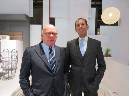 Zwei elegant gekleidete Männer im Anzug stehen lächelnd nebeneinander in einem modern gestalteten, hellen Büro- oder Messeraum mit Design-Deko im Hintergrund