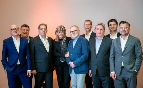 Neun geschäftlich gekleidete Personen posieren lächelnd nebeneinander vor neutralem Hintergrund in einem Konferenzraum