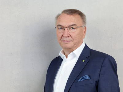 Älterer Mann mit Brille im dunkelblauen Anzug und weißem Hemd steht vor einer hellgrauen Wand und blickt ernst in die Kamera.