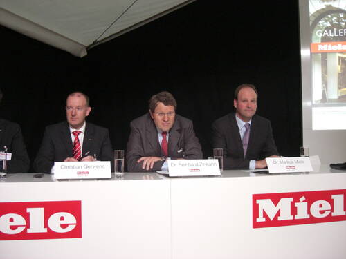 Drei Manager im Anzug sitzen nebeneinander hinter einem weißen Podium mit Miele-Logo und Namensschildern und sprechen auf einer Pressekonferenz vor dunklem Hintergrund.
