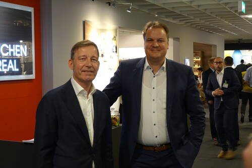 Zwei Männer in Anzügen posieren lächelnd in einer modernen Ausstellungs- oder Messehalle, während weitere Geschäftsleute im Hintergrund Gespräche führen.