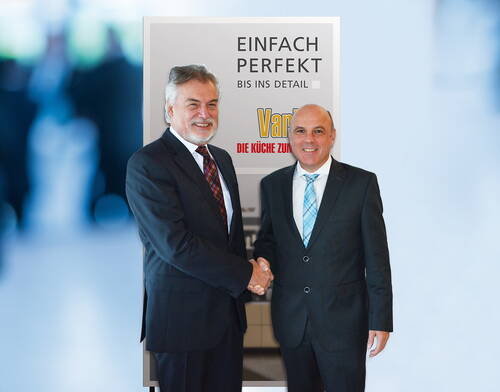 Zwei Geschäftsleute im Anzug geben sich vor einem Werbeplakat mit der Aufschrift „Einfach perfekt bis ins Detail“ lächelnd die Hand