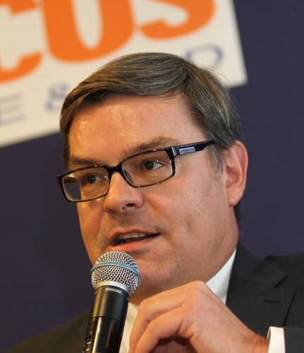 Mittelalterlicher Mann mit Brille spricht konzentriert in ein Mikrofon vor einem unscharfen Werbeplakat im Hintergrund