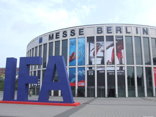 Große blaue IFA-Skulptur vor dem Messausgang der Messe Berlin mit hohen Glasfassaden und bunten Werbebannern für Elektronikmarken