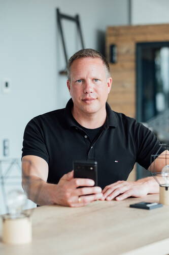 Mittelalterlicher Mann in schwarzem Polohemd sitzt an einem Holztisch und blickt mit einem Smartphone in der Hand direkt in die Kamera in einem modern eingerichteten Büro.