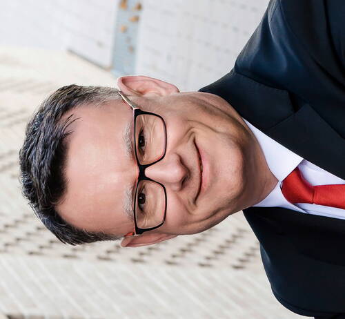 Mittelalterlicher Mann mit Brille, Anzug und roter Krawatte lächelt in die Kamera vor unscharfem, hellen Hintergrund.