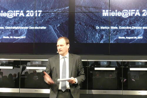 Geschäftsmann im Anzug spricht mit Notizen in der Hand auf der Bühne der Präsentation „Miele@IFA 2017“ vor einer Wand aus Einbauöfen