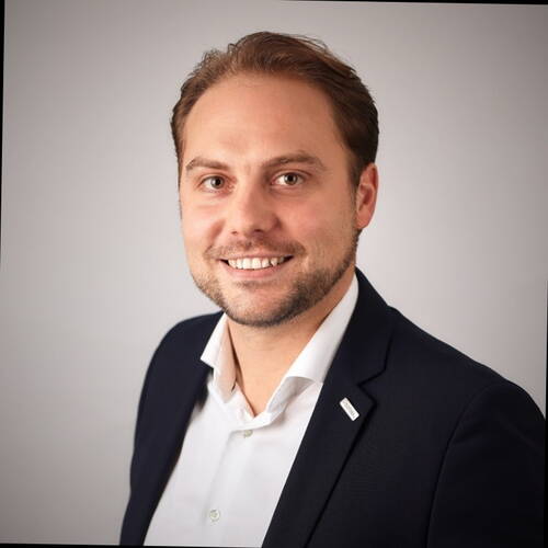 Lächelnder Mann im Business-Anzug vor neutralem Hintergrund, leicht schräg zur Kamera gedreht