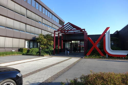 Eingangsbereich eines modernen Bürogebäudes mit rotem Stahlportal, großem XL-Skulpturlogo und Personen, die das Glasfoyer verlassen