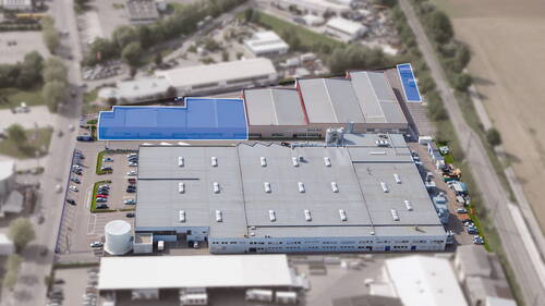Großes Industriegebäude mit blau markierten Hallenbereichen, umgeben von Parkplätzen, Straßen und Bahntrasse in einem Gewerbegebiet aus der Luftperspektive