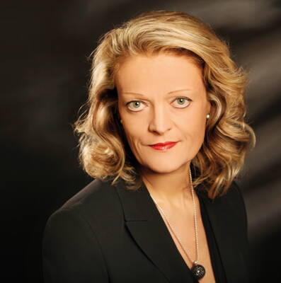 Elegant gekleidete Frau mit blond gelocktem Haar und rotem Lippenstift blickt ernst in die Kamera vor dunklem Hintergrund.