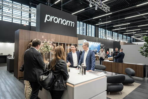 Geschäftsleute unterhalten sich an einem modernen Kücheninsel-Stand des Herstellers pronorm auf einer hell beleuchteten Messehalle.