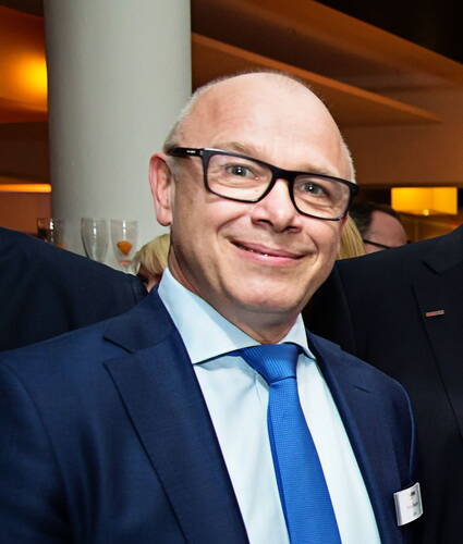 Glücklich lächelnder, glatzköpfiger Mann mit Brille im blauen Anzug und hellblauer Krawatte auf einer festlichen Abendveranstaltung in Innenräumen