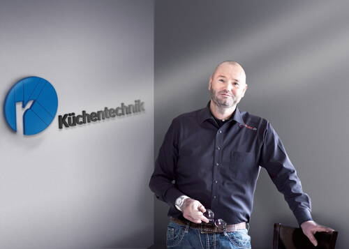 Mann in dunklem Hemd lehnt lässig an einem Stuhl in modernem Büro vor grauer Wand mit blauem „Küchentechnik“-Logo.