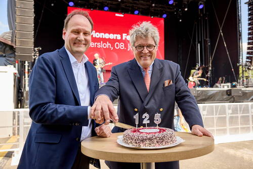Zwei festlich gekleidete Männer schneiden gemeinsam eine Jubiläumstorte mit der Zahl 125 vor einer Bühne bei einer Open-Air-Veranstaltung an.